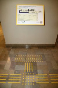Plan wystawy Zdjęcie dotykowego planu piętra wystawy, z napisami w alfabecie Brajla. Pod nim żółte ścieżki naprowadzajace na tle kamiennej podłogi.