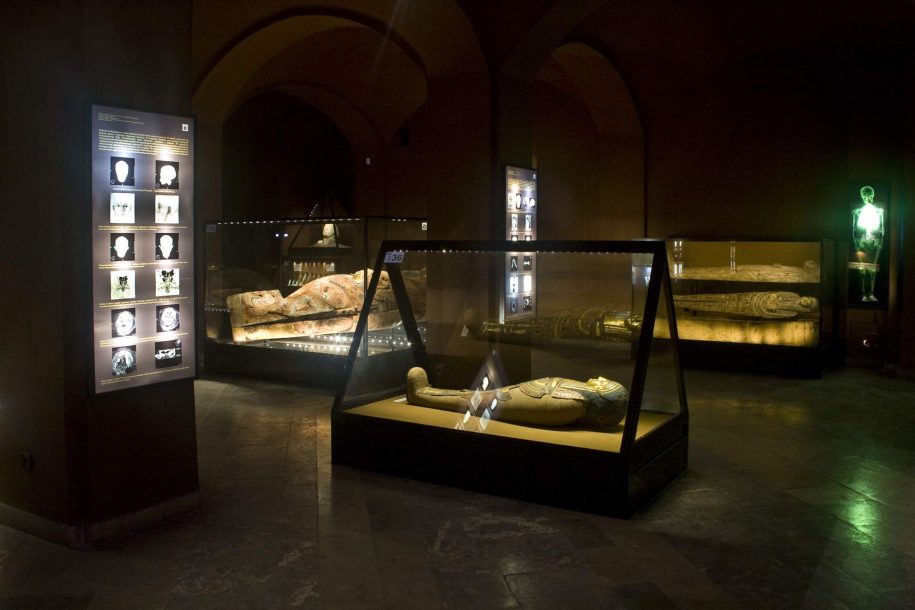 Fotografia kolorowa. W ciemnej sali wystawowej stoją trzy duże gabloty. W dwóch znajdują się sarkofagi z mumią, w trzecim model mumii z wyeksponowaną na nim starożytną ozdobą wykonaną z malowanych bandaży. Na dwóch filarach wiszą dwie prostokątne, podświetlane tablice zawierające zdjęcia z badań mumii i ich opisy.