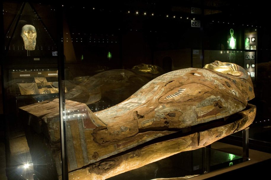 Zdjęcie kolorowe. Drewniany egipski sarkofag (trumna) w sali ekspozycyjnej. Sarkofag z wyglądu przypomina człowieka. Ma złotą twarz i otwarte, czarne oczy. Na tułowiu pokrytym niewyraźnymi malowidłami widać pęknięcia i ubytki. Na drugim planie egipska maska ze złotą twarzą.