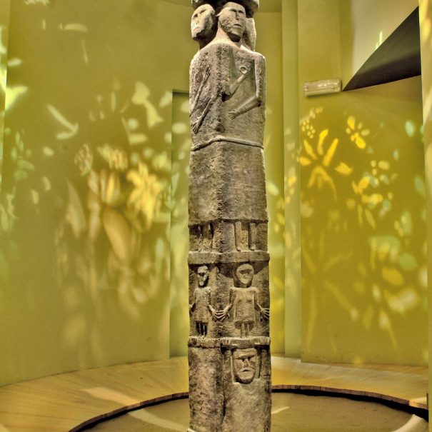 Zdjęcie kolorowe. Sala wystawowa z kamiennym posągiem w kształcie czworobocznego słupa. Górna część posągu to kapelusz, pod którym znajdują się dwie twarze bogów; widać również fragment profilu trzeciej twarzy. Poniżej, do połowy wysokości słupa, przedstawione są postacie ludzi trzymające się za ręce. W dolnej części posągu wyobrażenie klęczącej postaci podtrzymującej wyższe rzędy.