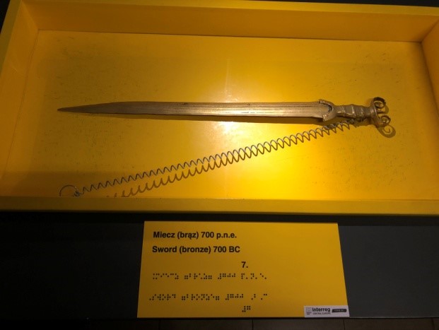 Kopia zabytkowego miecza W żółtym pojemniku krótki miecz z brązu. Podpis w czarnodruku i brajlu w językach polskim i angielskim.