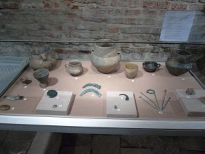 Kolorowe zdjęcie z wystawy. Przedstawia szklaną gablotę, w której widać gliniane naczynia różnej wielkości oraz metalowe narzędzia. Za gablotą widać fragment ceglanej ściany.