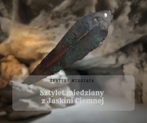 Grafika, zdjęcie frontalne sztyletu miedzianego z dwoma otworami na nity. Częściowo pokrytego zieloną patyną. Z małą plamą oczyszczonej powierzchni – lśniącej miedzi. W tle ciemna jaskinia lekko doświetlona słońcem. Z podpisem: Zabytek Miesiąca, Sztylet miedziany z Jaskini Ciemnej.