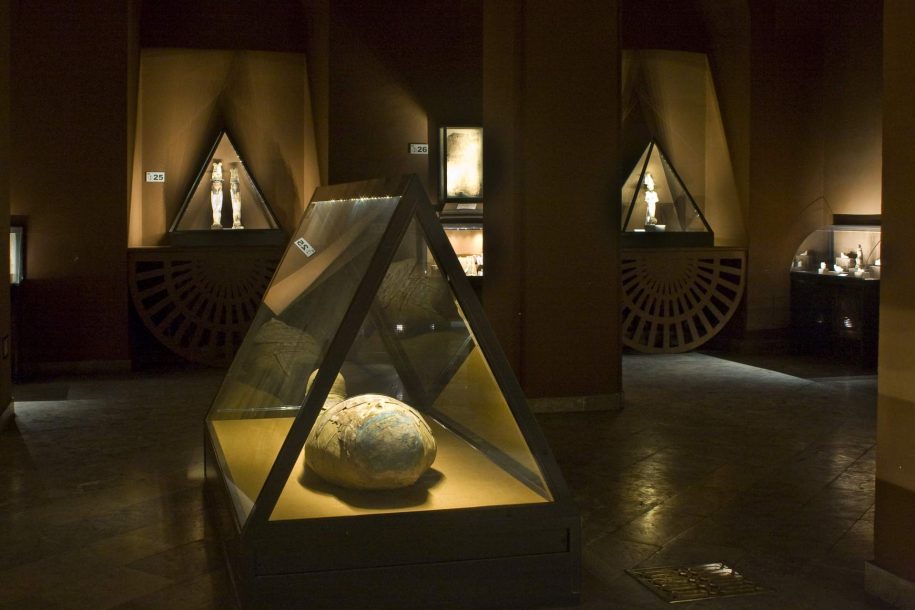 Zdjęcie kolorowe. Wnętrze sali wystawowej. W tle widać niewielkie trójkątne gabloty z drewnianymi figurkami. Na środku sali duża trójkątna gablota z modelem mumii przykrytym kartonażem – utwardzoną, zdobioną tkaniną.