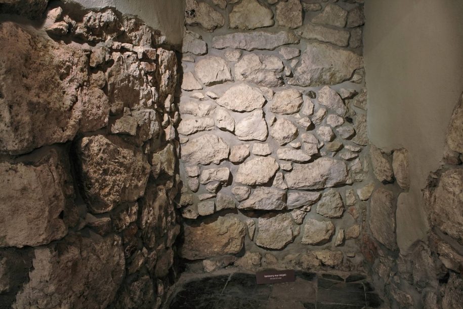 Fotografia kolorowa. Nisza w murze. Na wprost i po bokach znajdują się trzy ściany zbudowane z kamienia i zaprawy. Górne części bocznych ścian pokryte są zaprawą. Podłogę tworzą czarne płytki.