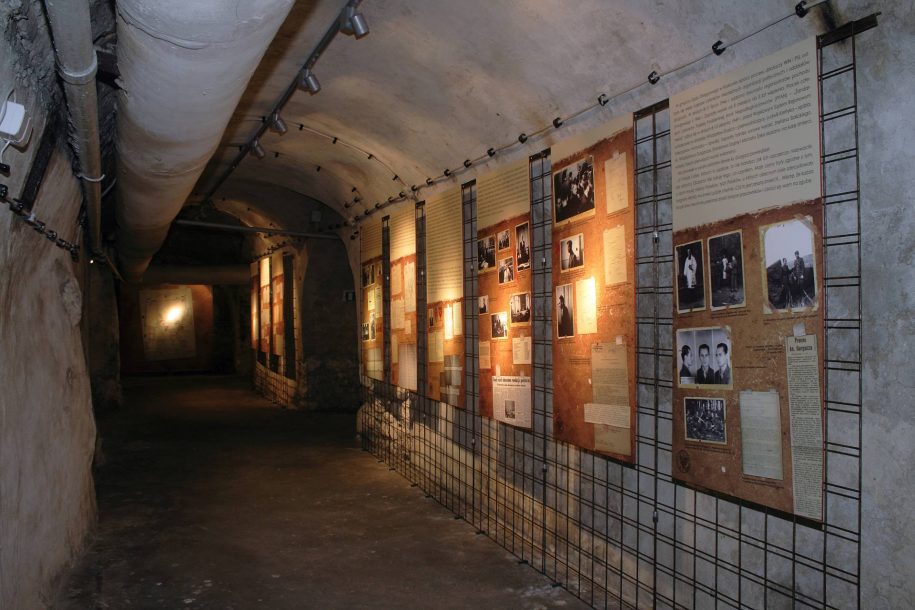 Fotografia kolorowa. Długi, wąski tunel, na którego końcu znajduje się tablica z odbijającą się w niej plamą światła. Z lewej strony, pod sufitem ciągną się rury. Z prawej strony, na ścianie wiszą tablice informacyjne z tekstem i zdjęciami przedstawiającymi ludzi.