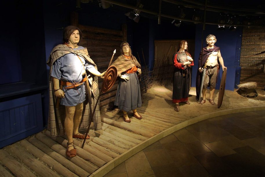 Zdjęcie kolorowe. Sala wystawowa. W półokręgu stoją cztery manekiny ubrane w stroje historyczne: mężczyzna, dwie kobiety i mężczyzna. Ubrane są w kolorowe repliki pradziejowych strojów.
